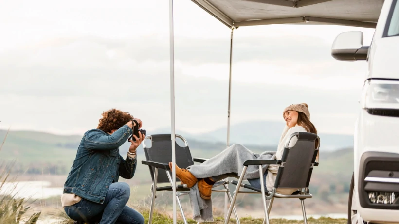 A man photographs a woman seated by a camper van A man photographs a woman seated by a camper van, capturing their joyful moment in nature during a road trip.